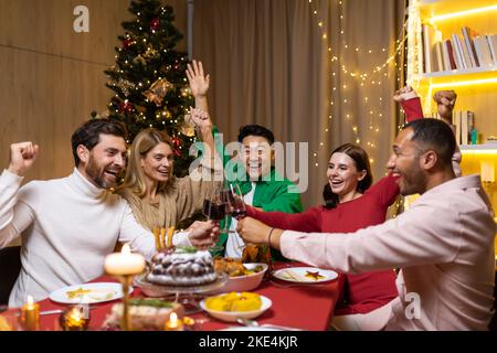 Young cheerful group of interracial people celebrating the coming new year. They sit at the festive table, clink glasses, raise their hands, congratulate each other, laugh, rejoice. Stock Photo