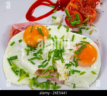 Top view on two fried eggs sunny side yellow runny yolk up on brad with ham, tomatoes and green fresh chives Stock Photo