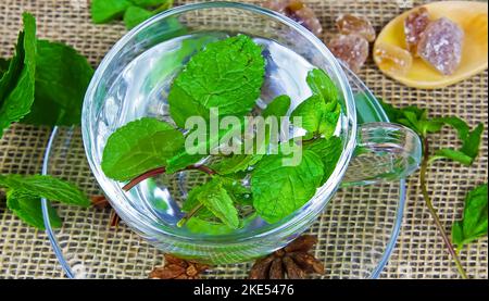 Closeup of one isolated transparent tea glass cup with fresh mint leaves, wood spoon, brown candy rock sugar, jute mat background. Stock Photo