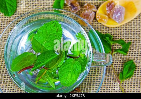 Closeup of one isolated transparent tea glass cup with fresh mint leaves, wood spoon, brown candy rock sugar, jute mat background. Stock Photo