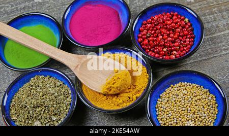 Top view on six blue isolated bowls with various colorful healthy spices, powder and grain condiments on wood table. Spoon in turmeric curcuma bowl. Stock Photo