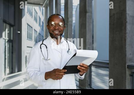 Portrait of senior doctor wearing glasses African American man looking at camera and smiling outside modern clinic, holding documents of patient's treatment report, satisfied with work result. Stock Photo