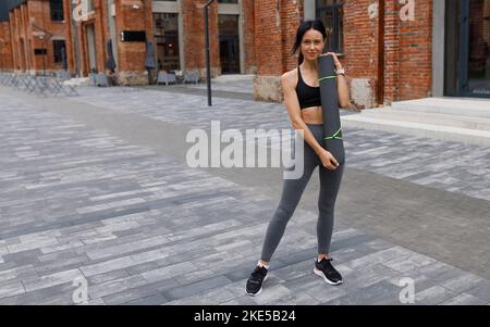 Strong athletic woman in stylish sportswear posing with yoga mat outdoors Stock Photo