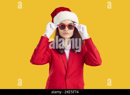 Woman in red suit, Christmas hat, white gloves and sunglasses isolated on yellow background Stock Photo