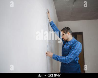 Side view of serious man using measuring tape in room with underfloor heating pipes. Designer in shirt taking measurements while working on renovation of apartment. Stock Photo