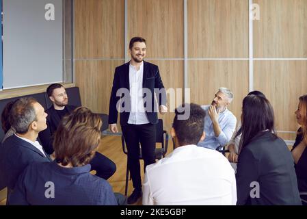 Cheerful businessman standing talking to colleagues who are sitting in circle around him in office. Stock Photo