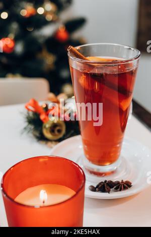 Close-up of candle and mulled red wine with aromatic spices and citrus fruits on white table on background decorated Xmas tree. Traditional hot drink Stock Photo
