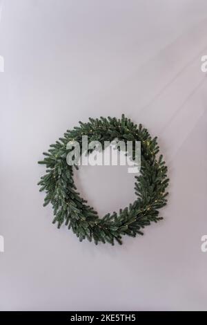 A green wreath of fir branches without Christmas toys hangs on a light wall. Handmade natural decor for the holiday. Home decoration in Rustic style. Stock Photo