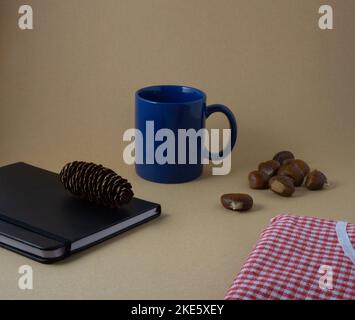 autumn composition with blue cup, black notebook with pine cone on top, chestnuts and red blanket Stock Photo