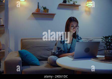 Tired focused woman using laptop at night, working overtime at home sitting on sofa, flexible working hours Stock Photo
