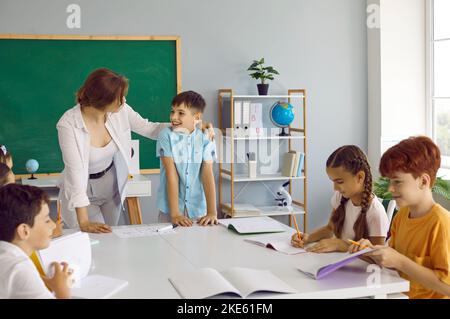 Friendly smiling teacher listens with pleasure to small schoolboy who answers at blackboard. Stock Photo