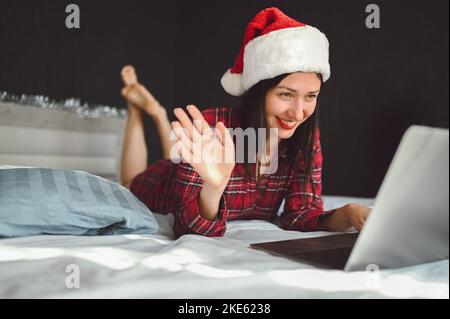 Excited smiling brunette woman in red pajamas lies in bed in New Year's Santa Claus hat waving have laptop video chat in dark interior bedroom. Young female at home in happy Christmas mood. Stock Photo