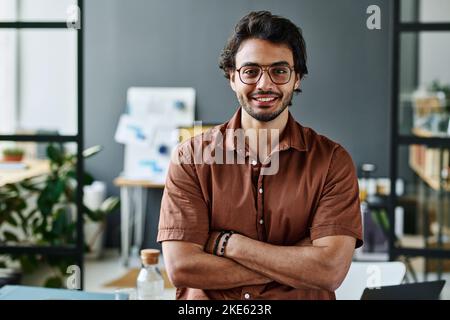 Young successful intern in eyeglasses and brown shirt keeping his arms crossed by chest while standing by workplace in front of camera Stock Photo