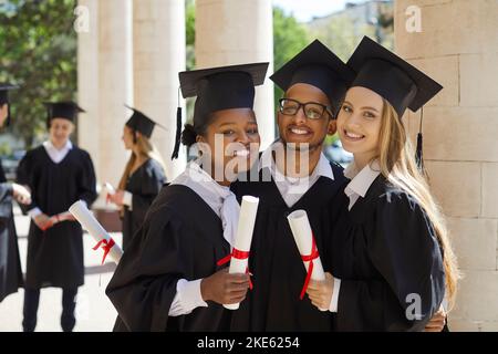 Portrait of three university graduate friends with diplomas in their hands outdoors after ceremony. Stock Photo