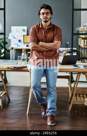 Young confident businessman in smart casualwear and eyeglasses crossing arms on chest while standing against his workplace Stock Photo