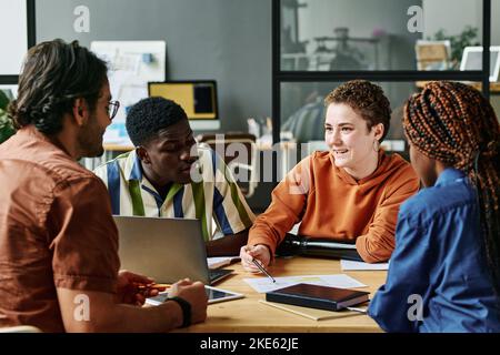 Young confident businesswoman with partial arm sharing her ideas with intercultural colleagues while sitting among them at meeting Stock Photo