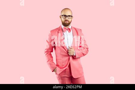 Bald man in pink suit and glasses posing with hand in pocket on pink studio background Stock Photo
