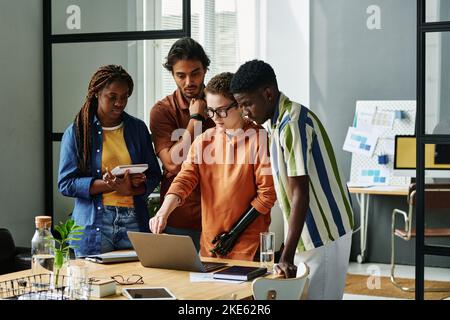 Young confident designer with partial arm pointing at laptop screen while making presentation of project plan to intercultural colleagues Stock Photo