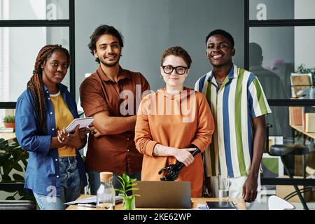Group of young creative designers in casualwear standing by workplace with laptop and supplies in office and looking at camera Stock Photo