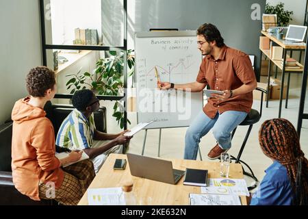 Young confident businessman pointing at graph on whiteboard and explaining it to colleagues listening to him and making notes Stock Photo