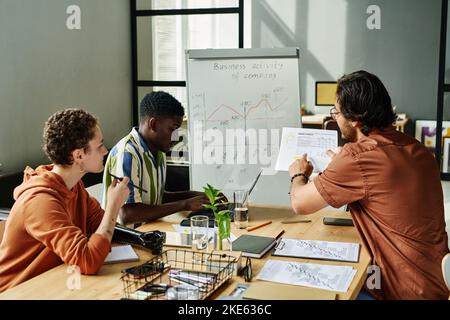 Young businessman pointing at financial document and explaining data to colleagues while one of them typing at working meeting Stock Photo
