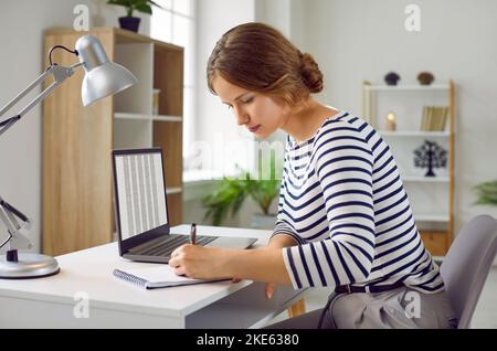 Left-handed woman makes notes in her notebook while working on laptop in office. Stock Photo