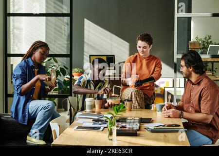 Group of young intercultural coworkers having snack and coffee for lunch in the middle of working day by workplace in office Stock Photo