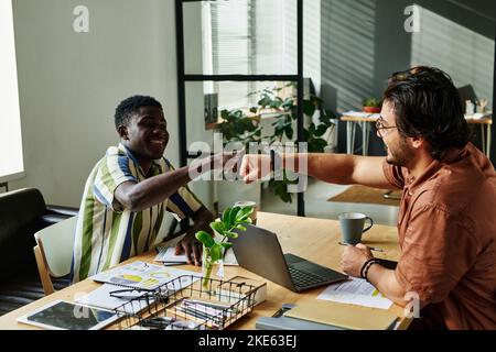 Two happy young intercultural colleagues making punch bump over workplace while congratulating one another on successful deal Stock Photo