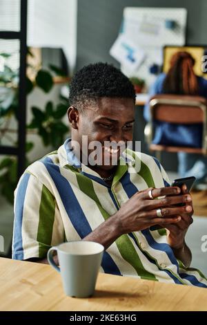 Happy young African American businessman texting in smartphone while sitting by workplace and having coffee at break in office Stock Photo
