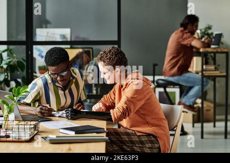 Two young intercultural interns discussing notes in copybook at working meeting while African American guy pointing at page Stock Photo