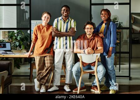 Group of young multicultural colleagues in casualwear standing against workplace in office and looking at camera with smiles Stock Photo