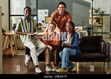 Four young successful interracial employees or creative designers in casualwear sitting on black leather couch in openspace office Stock Photo