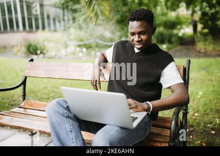Young black man in vest working on laptop computer outdoors, looking at screen, smiling. Student sitting on bench in college campus, enjoying freelance remote Job or distance education Stock Photo