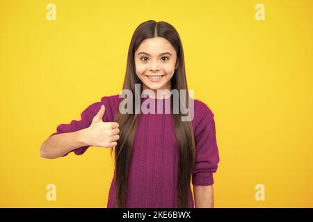 Like it. Portrait of joyful teenage child girl showing thumbs up and smiling, yellow background with copy space. Stock Photo