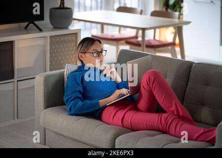 Young woman lying on comfortable sofa works on laptop and types information about business Stock Photo