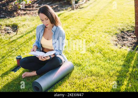 Pretty sporty woman making notes in a diary sitting on a sunny lawn in a summer park. Copy space. Green nature. Stock Photo