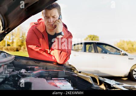 A male driver stands near the engine compartment of the car, calls the phone - asks for help. Transport concept. Stock Photo