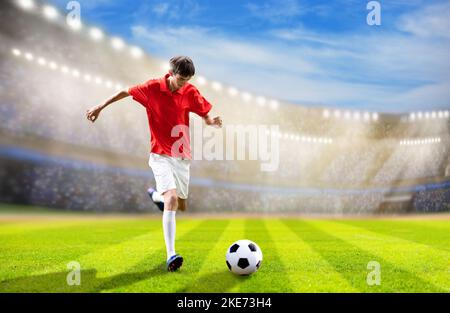 Football player on stadium field. Soccer match. Young man in red shirt and white shorts kicking ball on outdoor pitch. Athlete running and jumping. Stock Photo