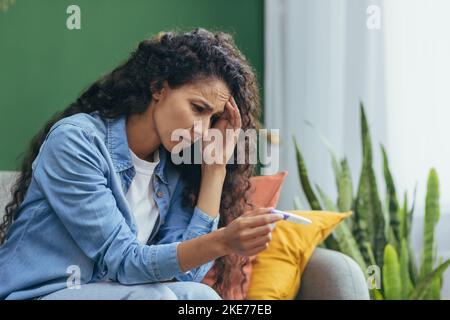 Sad and upset woman alone at home unhappy with pregnancy test, depressed hispanic woman sitting on sofa in living room. Stock Photo