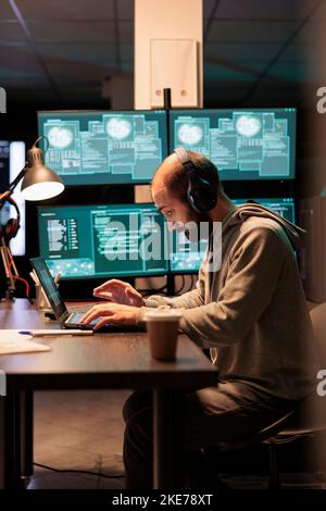 Cyber criminal trying to hack computer server, wearing headphones and working late at night. Hacker breaking into security system to steal information, hacking password and writing malware. Stock Photo