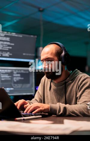 Young app developer writing code and listening to music on headphones, using artificial intelligence script to program big data on laptop. Programming server on terminal window. Stock Photo