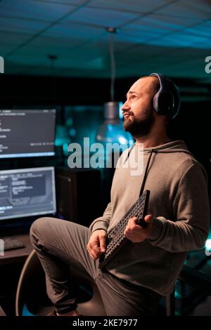 Male coder singing and playing guitar with keyboard, using IT equipment and fooling around. Silly funky programmer celebrating success, wokring on server code after hours in office. Stock Photo