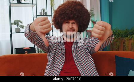 Dislike. Upset man showing thumbs down sign gesture, expressing discontent, disapproval, dissatisfied bad work at modern home apartment indoors. Displeased young guy in living room sitting on sofa Stock Photo