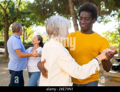 Exited older and middle-aged diverse people enjoying latin dances breaking into pairs on sandy area between trees Stock Photo