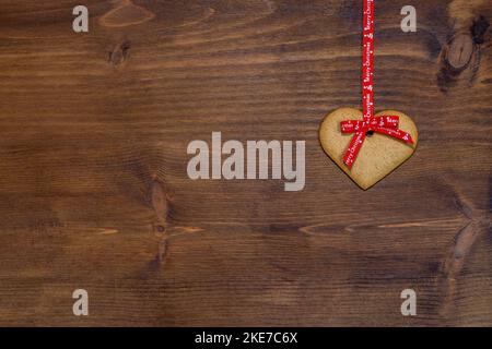 Heart-shaped gingerbread biscuit, typical for Christmas, hanging with a red ribbon on a wooden background. Stock Photo