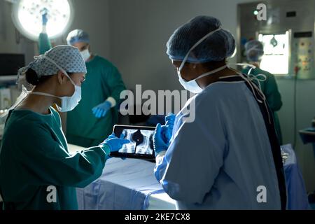 Two diverse female surgeons preparing for operation, studying x-ray on tablet in operating theatre Stock Photo