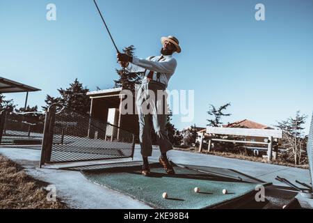 A wide-angle shot of an elegant bearded mature black man in a straw hat and trousers with suspenders, hitting the ball using a club during a golf trai Stock Photo