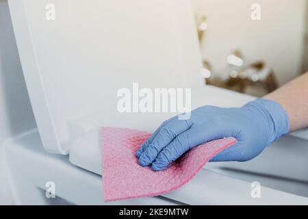 Close up on woman hand gloved in blue rubber protective gloves. Housewife cleaning toilet bowl, seat with detergent liquid, wet wipe in bathroom, restroom. Female washing, disinfecting toilet seat Stock Photo
