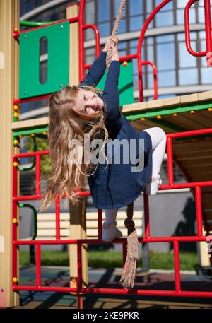 Adorable blond female child playing on climbing frames, smiling in sunny day. Cheerful girl climbing, holding hands, hanging upside down at playground. Concept of kids activities. Stock Photo