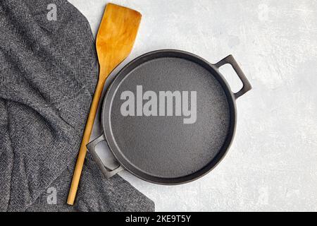 Empty black iron empty frying pan, wooden spatula and gray kitchen towel on a gray concrete background Stock Photo